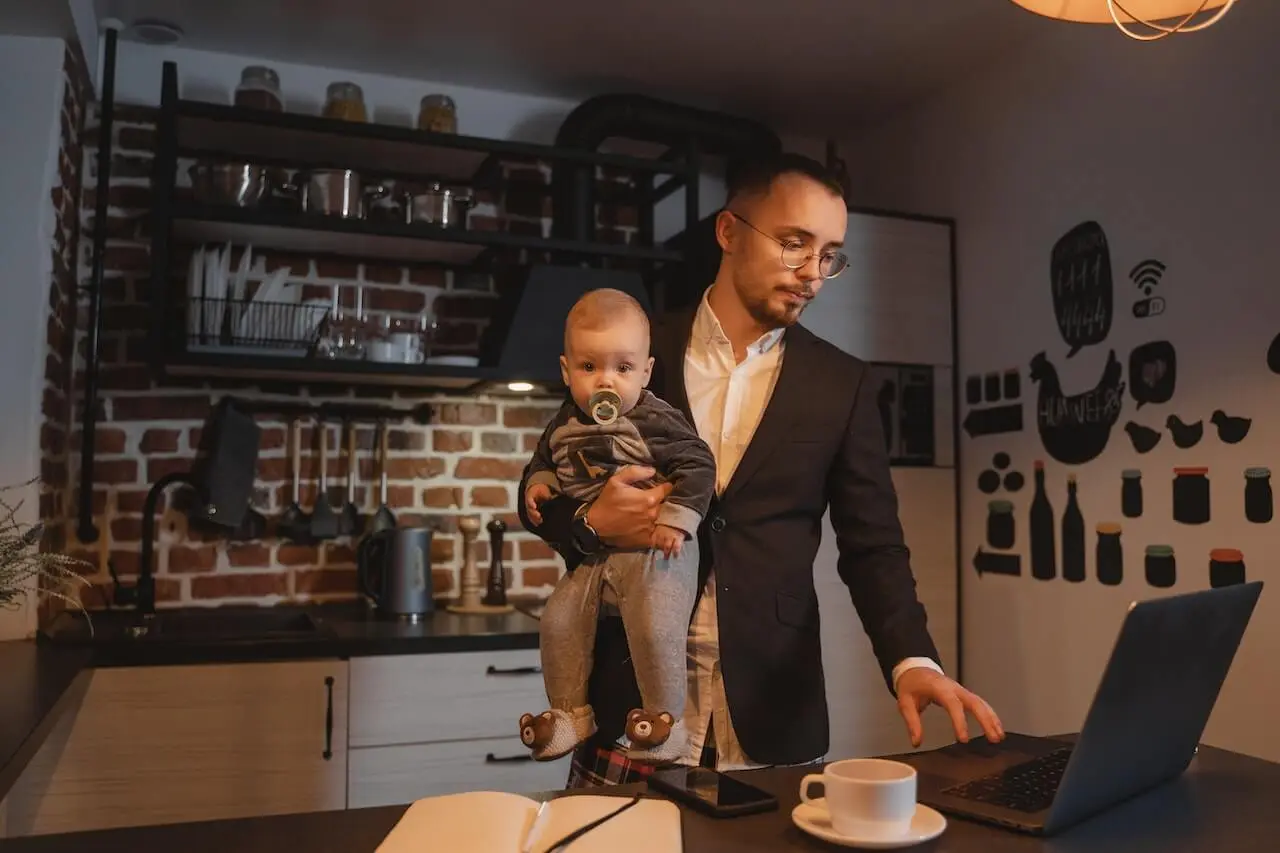 A man works at a laptop as a remote worker while holding a baby