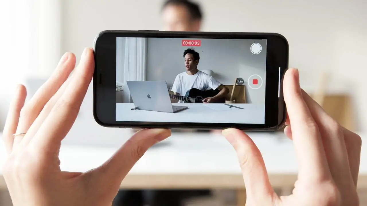 Image of a person holding a phone recording a video of someone with a Macbook and a guitar.