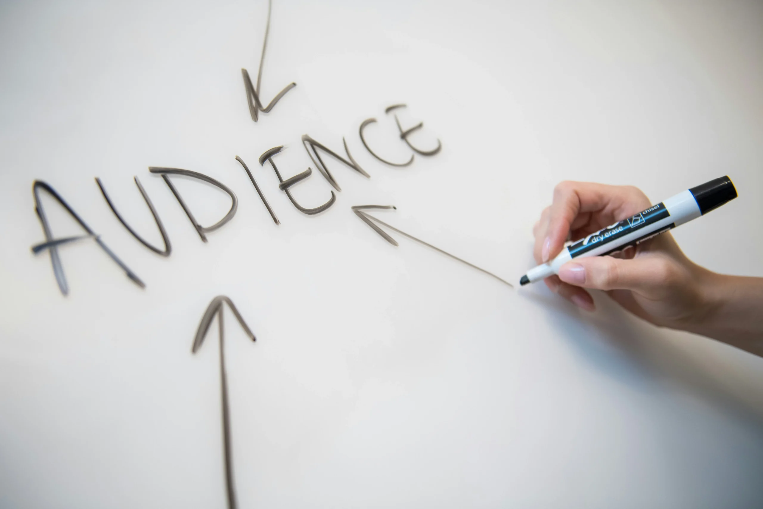Close-up of a hand using a black dry erase marker to draw arrows pointing towards the word AUDIENCE on a white board.
